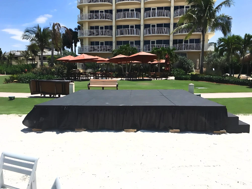 An outdoor stage set up on the sand, surrounded by grass, with a black skirt and wooden supports, under a bright sunny sky.